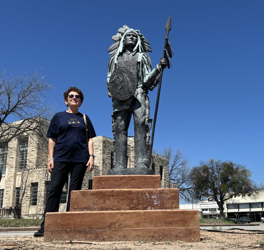 A person stands beside a large bronze statue of a Native American warrior holding a spear and shield against a clear blue sk…