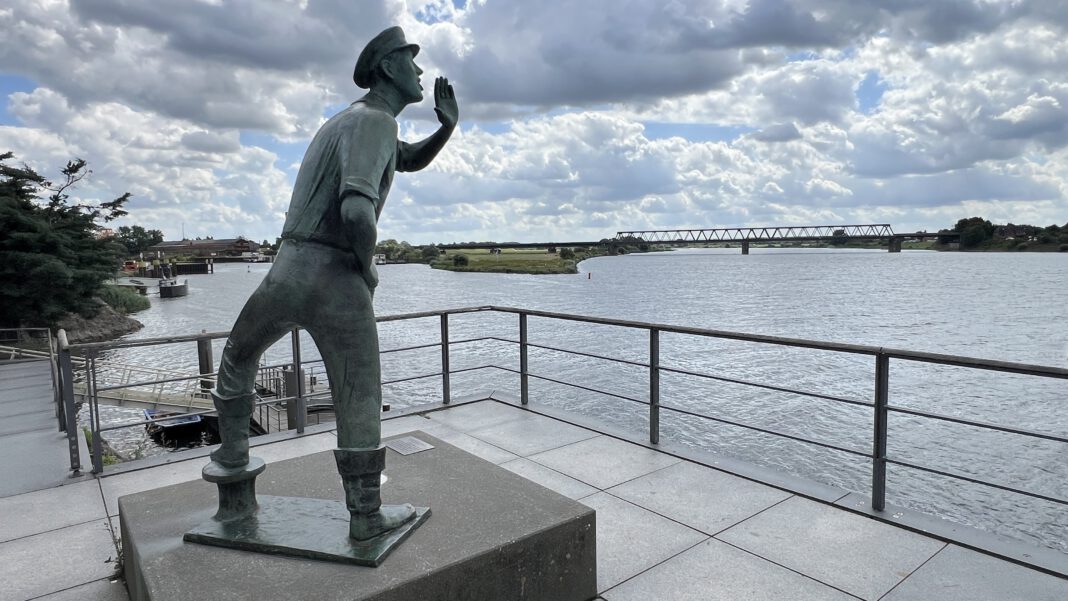A bronze statue of a sailor gesturing toward the river under a partly cloudy sky, with a bridge visible in the background.